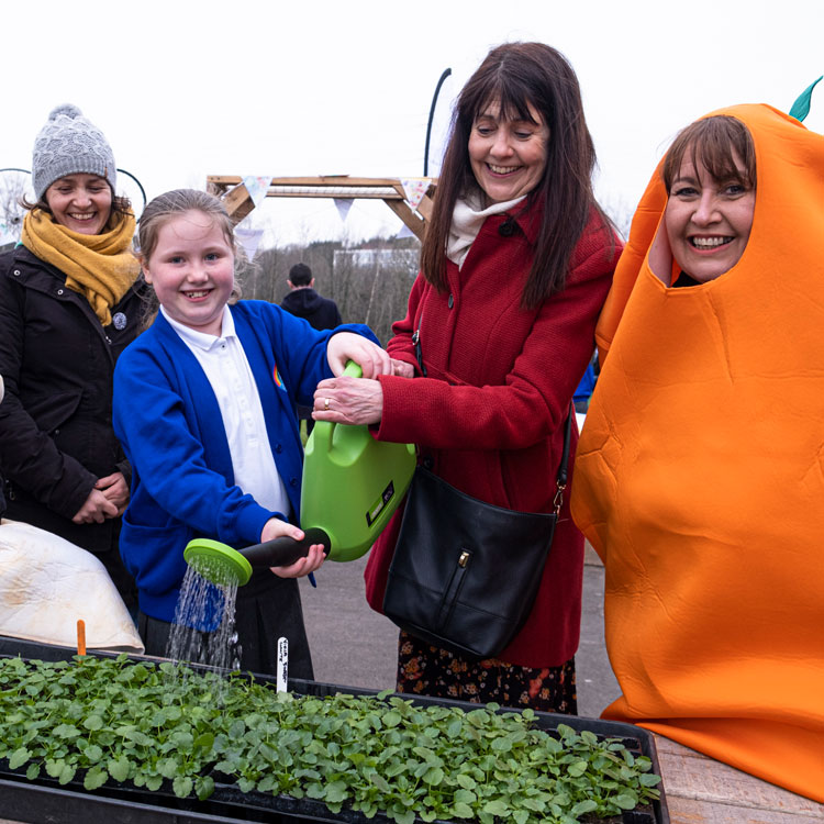 Child & three adults watering plants in edible playground. One of the adults is dressed as a carrot