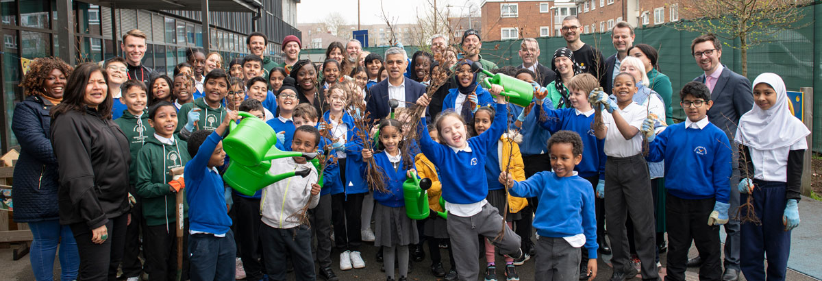 Group of smiling children & adults in a school yard holding saplings & watering cans