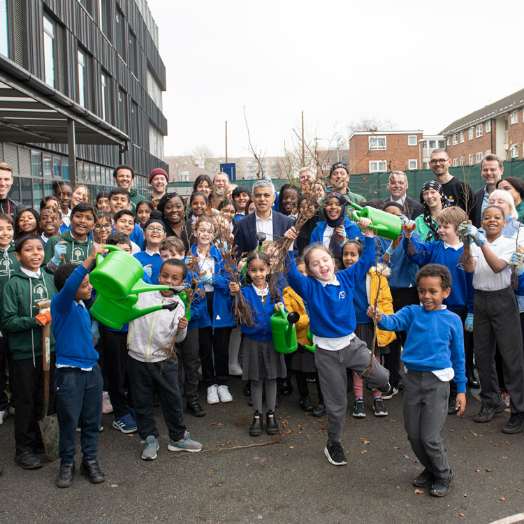 Group of smiling children & adults in a school yard holding saplings & watering cans