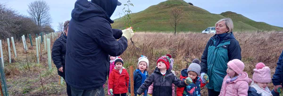 Young children watching an adult plant a tree next to steep hills