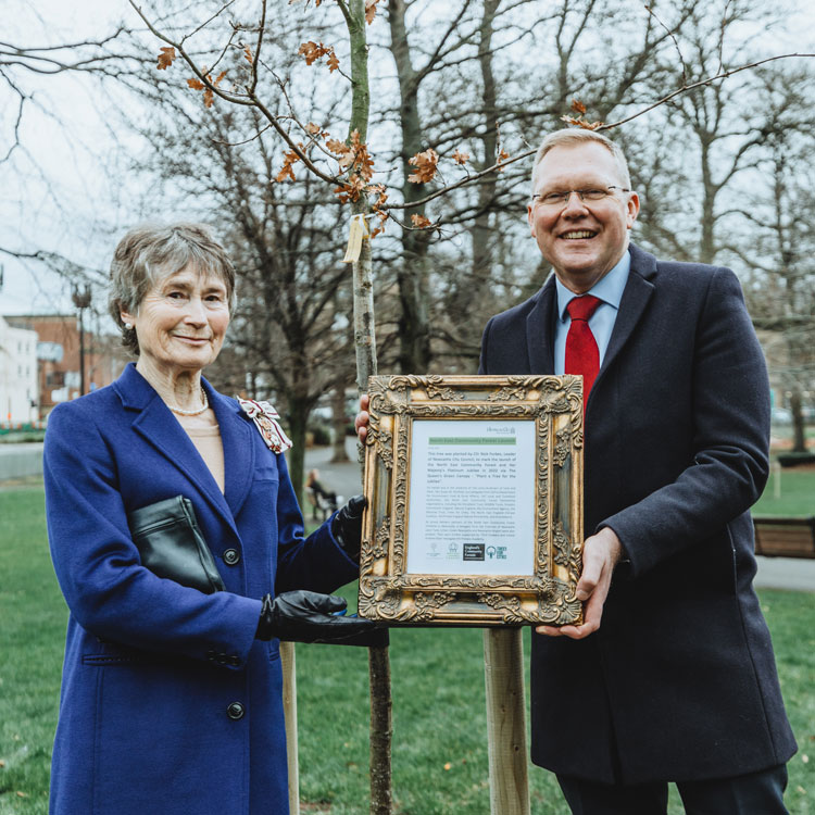 Two adults standing infront of a tree holding a certificate